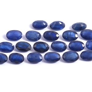May include: A collection of oval-cut, deep blue sapphires arranged on a white surface. The gemstones exhibit a polished, faceted appearance, reflecting light and showcasing their rich color. The sapphires are of varying sizes and are ideal for jewelry making.