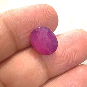 May include: A vibrant, oval-shaped, translucent magenta-pink gemstone with a faceted surface. The stone displays internal inclusions and varying shades of deep pink and purple throughout its structure, reflecting light across its polished surface.