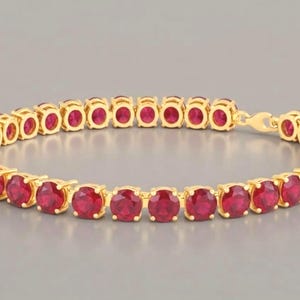 Puede incluir: Una pulsera de tenis de oro con una fila continua de rubíes redondos de color rojo vibrante engastados en garras de oro individuales. La pulsera está dispuesta en forma circular, mostrando un acabado de metal dorado pulido y un cierre seguro.
