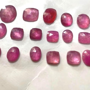 May include: A collection of sixteen loose, faceted gemstones in various shades of pink and red. The stones are arranged in three rows and feature a mix of oval, round, and cushion shapes with polished, reflective surfaces and visible geometric facets.