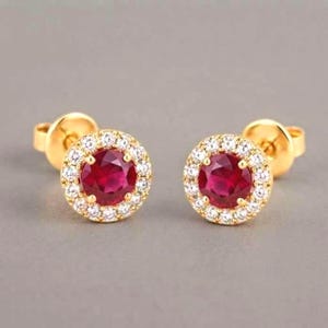 May include: A pair of gold stud earrings featuring a round, deep red center stone surrounded by a halo of small, clear, sparkling white stones. The earrings have a classic post-back closure and a polished yellow gold metal setting.