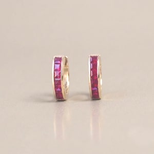 May include: A pair of gold hoop earrings featuring a channel setting with multiple square-cut vibrant red rubies. The earrings have a polished metallic finish and a hinged closure mechanism, showcasing a sleek and uniform design throughout.