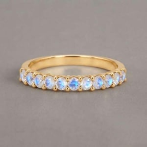 May include: A gold band ring featuring a continuous row of round, faceted moonstones. Each stone displays a soft, iridescent blue glow set within a polished gold metal setting that encircles the entire circumference of the ring.