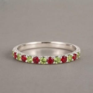 Può includere: Un anello a fascia in argento con una fila di pietre preziose rotonde alternate in rosso e verde chiaro, fissate con una montatura a griffe condivisa. L'anello presenta una finitura metallica lucida e un profilo sottile con motivo.