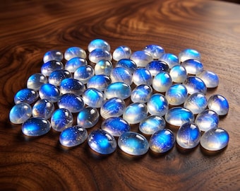 May include: A collection of oval-shaped, polished moonstone cabochons. Each stone displays a captivating blue adularescence, with a milky, iridescent glow. The gemstones are scattered on a dark, wooden surface.