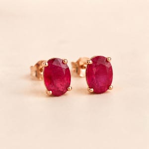 Pode incluir: Um par de brincos de tarracha com rubis vermelhos vibrantes de corte oval cravados em ouro rosa. Cada pedra preciosa é fixada por quatro pequenas garras, exibindo um tom carmesim profundo e uma superfície polida e facetada que reflete a luz.