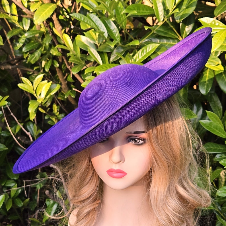 Puede incluir: Un sombrero morado vibrante con ala ancha y copa redonda. El sombrero est&aacute; hecho de material tejido y tiene una forma estructurada. El sombrero se encuentra sobre un fondo de follaje verde.