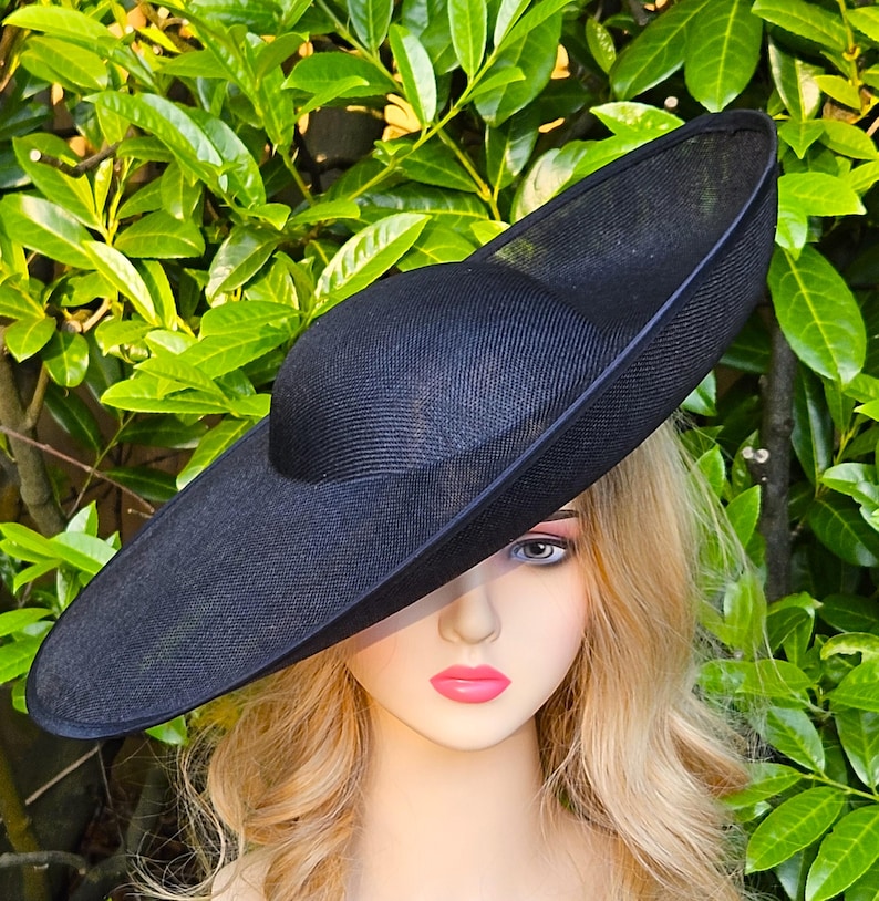Puede incluir: Un sombrero negro con ala ancha y copa redondeada. El sombrero est&aacute; hecho de un material tejido y tiene un aspecto ligeramente texturizado. El sombrero se encuentra sobre un fondo de follaje verde.