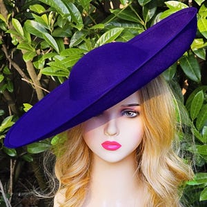Puede incluir: Un sombrero morado vibrante con ala ancha. El sombrero est&aacute; hecho de una tela texturizada y tiene una copa redondeada. El sombrero se coloca sobre un fondo de follaje verde.