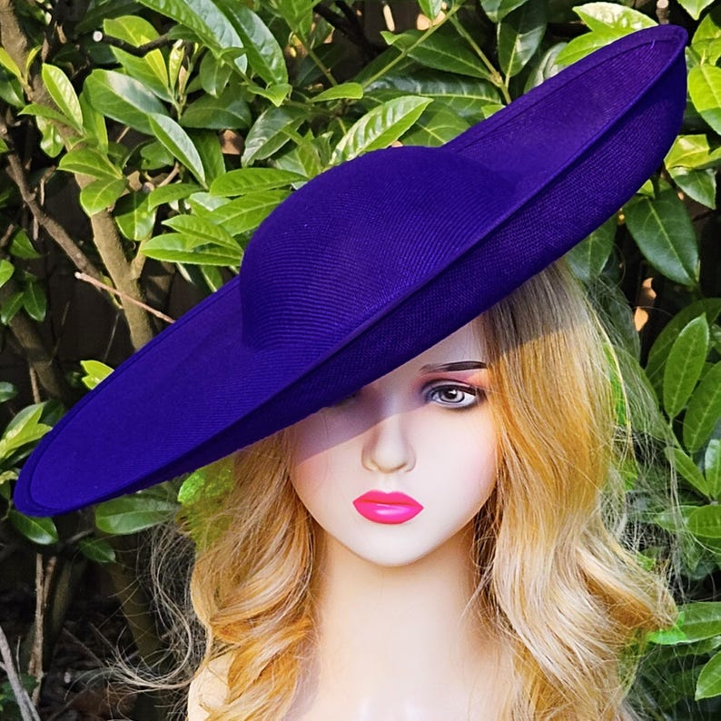Puede incluir: Un sombrero morado vibrante con ala ancha. El sombrero est&aacute; hecho de una tela texturizada y tiene una copa redondeada. El sombrero se coloca sobre un fondo de follaje verde.
