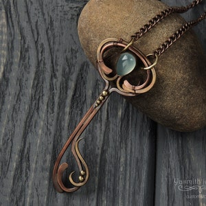 May include: A copper and brass key pendant necklace with a light blue gemstone. The necklace is on a chain and is resting on a brown rock.