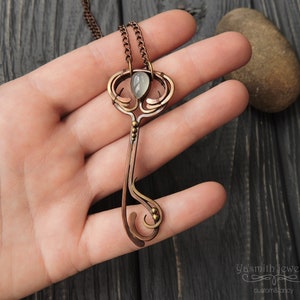 May include: A copper key pendant necklace featuring a green gemstone set in the top. The key has an intricate design with a swirl at the bottom and a small ball at the end of the handle. The necklace is on a chain with a silver finish.