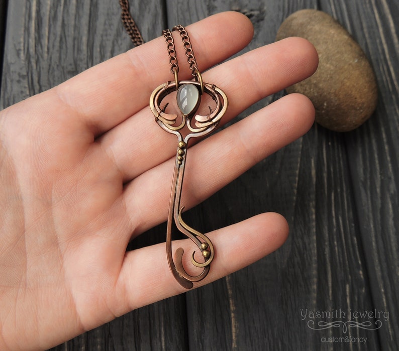 May include: A copper key pendant necklace featuring a green gemstone set in the top. The key has an intricate design with a swirl at the bottom and a small ball at the end of the handle. The necklace is on a chain with a silver finish.