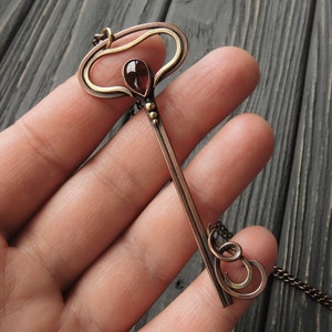 May include: A copper and gold key pendant necklace with a red gemstone. The key has a decorative top and a long, thin shaft. The necklace is on a chain.