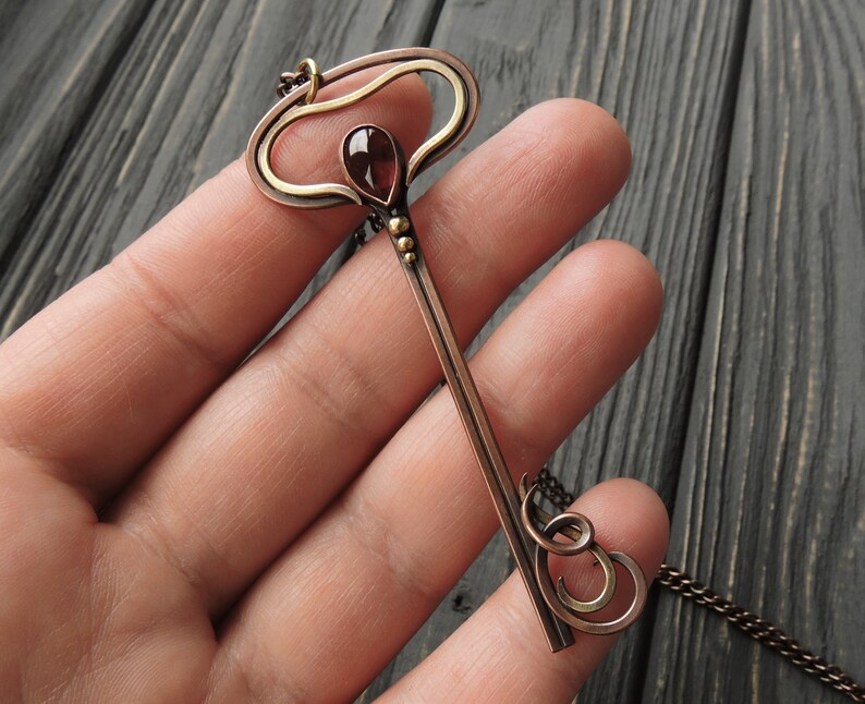 May include: A copper and gold key pendant necklace with a red gemstone. The key has a decorative top and a long, thin shaft. The necklace is on a chain.