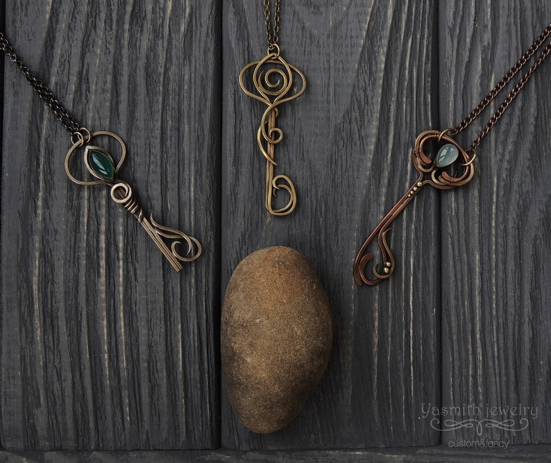May include: Three ornate key-shaped pendants with green and blue gemstones hanging from chains on a dark wood background. The pendants are made of wire and have intricate designs. The text "Yasmirh jewelry custom & fancy" is visible in the bottom right corner of the image.