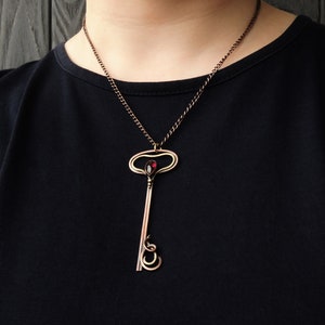 May include: A copper key pendant necklace with a red gemstone in the center. The key has a decorative, ornate design and is suspended from a copper chain.