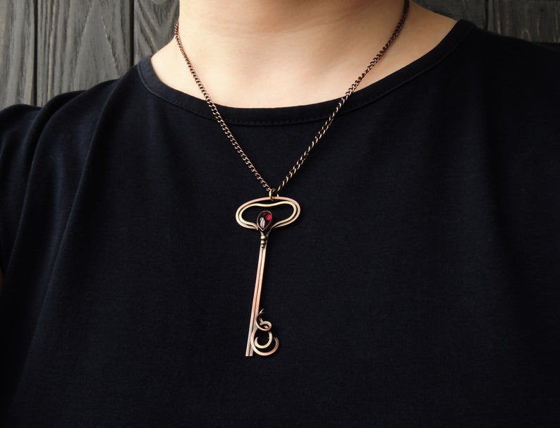 May include: A copper key pendant necklace with a red gemstone in the center. The key has a decorative, ornate design and is suspended from a copper chain.