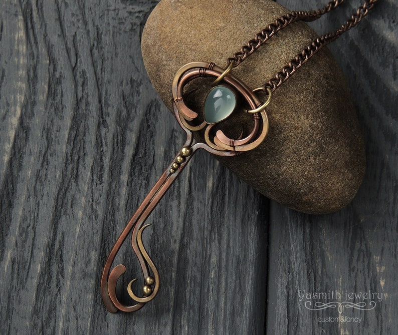 May include: A copper and brass key pendant necklace with a light blue gemstone. The necklace is on a chain and is resting on a brown rock.