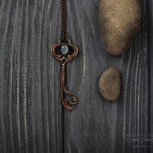 May include: A copper key pendant necklace with a light blue gemstone and gold accents. The necklace is on a dark wood background.