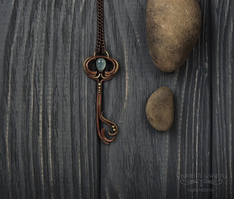 May include: A copper key pendant necklace with a light blue gemstone and gold accents. The necklace is on a dark wood background.