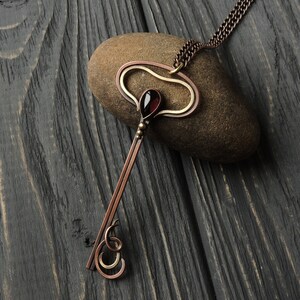 May include: A copper and gold key pendant necklace with a red gemstone. The necklace is on a dark wood background.