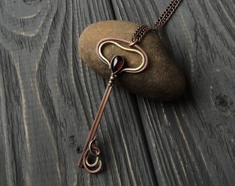 May include: A copper and gold key pendant necklace with a red gemstone. The necklace is on a dark wood background.