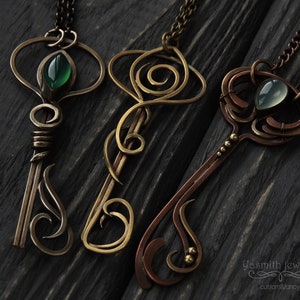 May include: Three ornate key-shaped pendants with green and blue gemstones. The pendants are made of copper and brass wire and have intricate designs. The pendants are hanging from chains on a dark wooden background.
