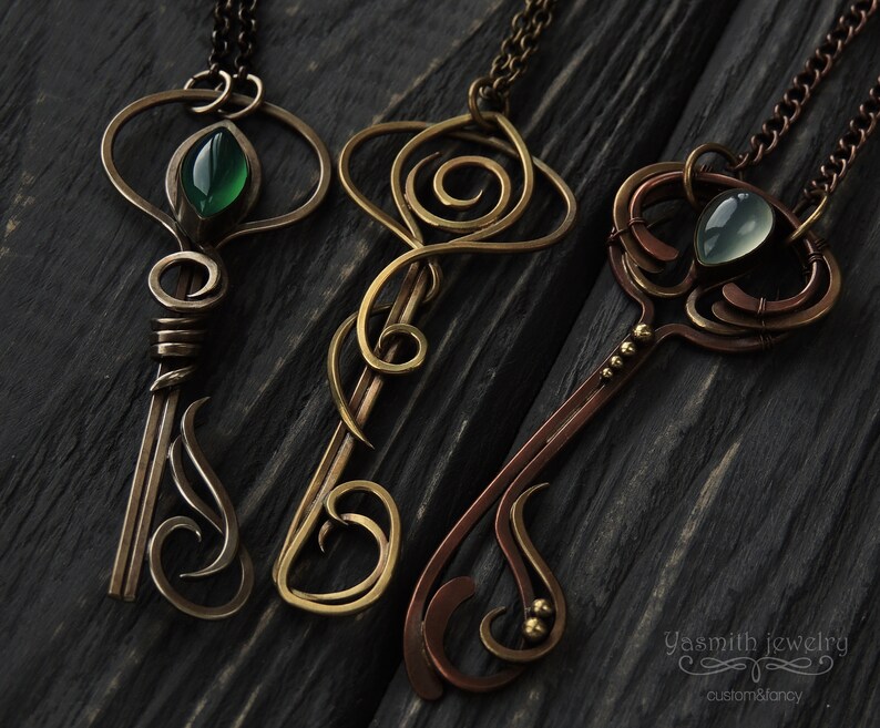 May include: Three ornate key-shaped pendants with green and blue gemstones. The pendants are made of copper and brass wire and have intricate designs. The pendants are hanging from chains on a dark wooden background.