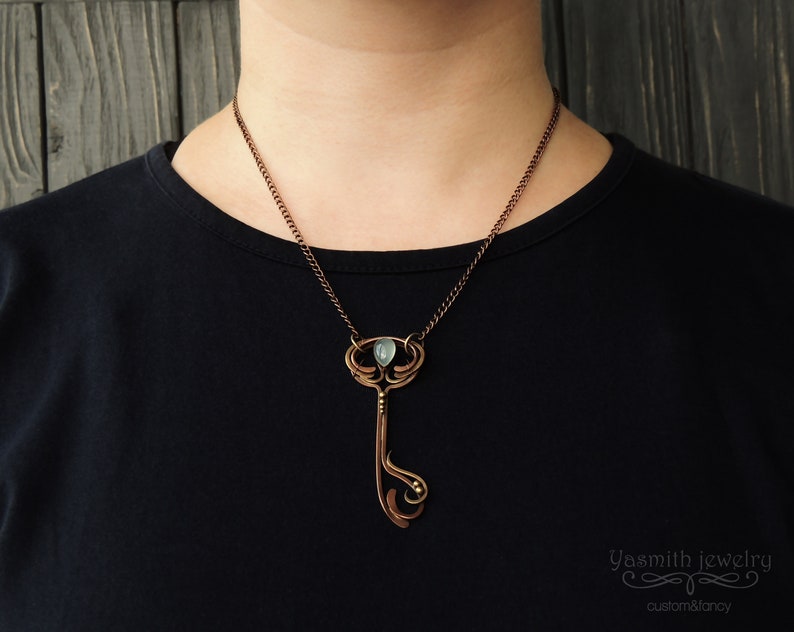 May include: A copper necklace with a key-shaped pendant featuring a green gemstone. The pendant has intricate details and a delicate chain.