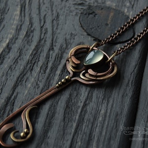 May include: A delicate copper and brass necklace with a light blue gemstone pendant in the shape of a key. The necklace features intricate wirework and a chain.