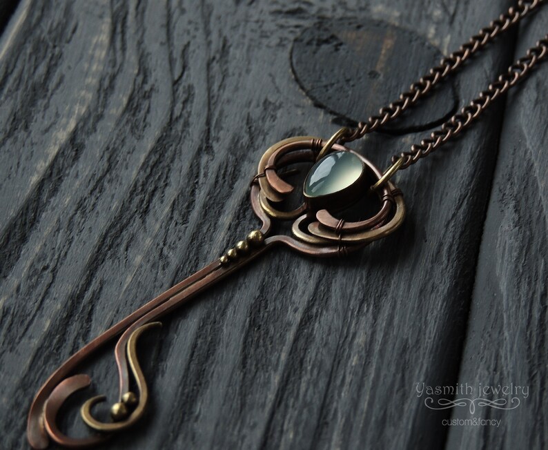 May include: A delicate copper and brass necklace with a light blue gemstone pendant in the shape of a key. The necklace features intricate wirework and a chain.