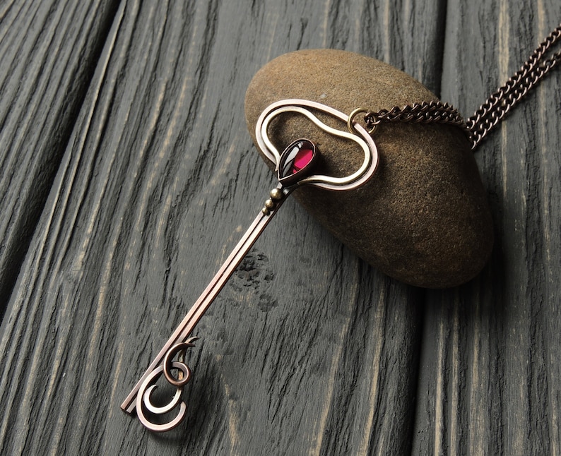 May include: A copper key pendant necklace with a red gemstone in the center of the keyhole. The necklace is on a brown wooden surface.
