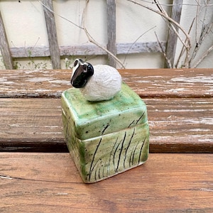 May include: A square ceramic lidded box with a green glaze and black painted grass details. A small, sculpted white sheep figurine with a black face and ears is attached to the top of the lid, serving as a handle for the container.