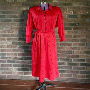 Puede incluir: Un vestido rojo con lunares negros, mangas largas y cinturón anudado. El vestido tiene cuello y se exhibe en un maniquí contra una pared de ladrillos. El vestido llega a la rodilla.