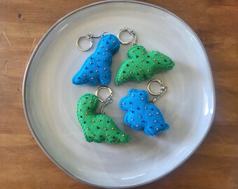 May include: Four handmade dinosaur keychains in blue and green felt, each with a silver keyring. The dinosaurs are decorated with small, gold-colored beads. The keychains are arranged on a light gray ceramic plate.