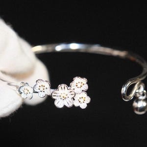 May include: A silver-toned bracelet featuring a floral design. The bracelet has a series of white flowers with gold-colored centers. A small silver ball dangles from the end of the bracelet. The bracelet is held by a gloved hand against a black background.