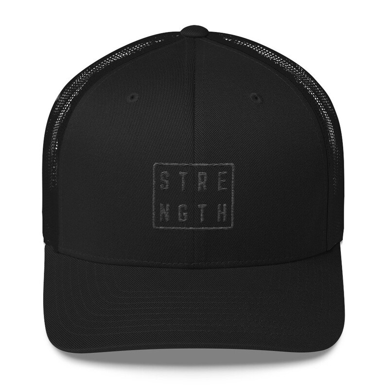 Puede incluir: Gorra de camionero negra con una parte trasera de malla negra y un parche cuadrado bordado en negro que dice "STRENGTH" en may&uacute;sculas.