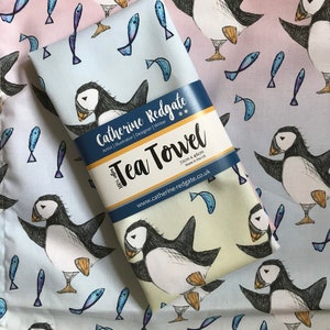 Könnte beinhalten: Ein weißes Geschirrtuch mit einem blauen und rosa Ombre-Design, das ein sich wiederholendes Muster aus Papageientauchern und Fischen zeigt. Der Text "Catherine Redgate" und "Tea Towel" ist auf das Handtuch gedruckt. Das Handtuch ist 72 cm x 46 cm groß und wurde in Großbritannien hergestellt.