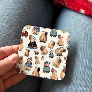 May include: A white square coaster with a repeating pattern of cartoon capybaras wearing different outfits. The capybaras are brown and white and are wearing sweaters, hats, and scarves.