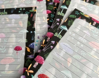 May include: A stack of lined notepads with a black background and a colorful mushroom pattern. The mushrooms are in various shades of pink, purple, blue, and green.