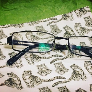 May include: Black rectangular-shaped eyeglasses with a black frame and clear lenses. The eyeglasses are lying on a white fabric with a repeating pattern of small gray and white animals.
