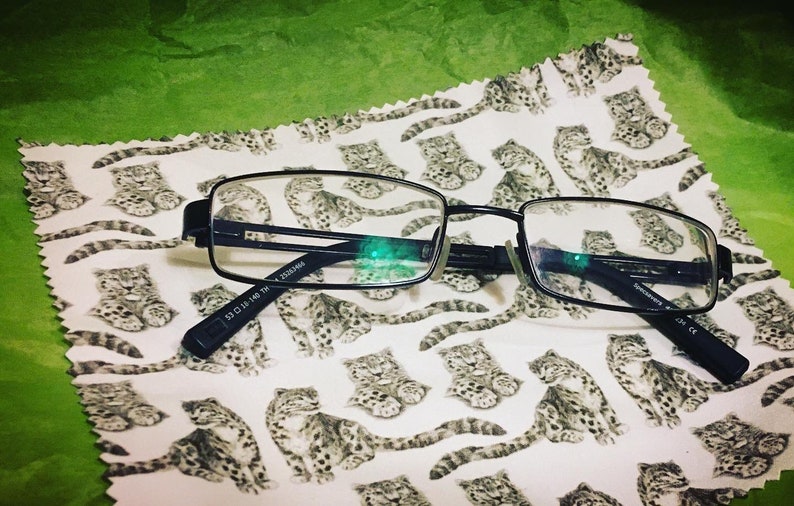 May include: Black rectangular-shaped eyeglasses with a black frame and clear lenses. The eyeglasses are lying on a white fabric with a repeating pattern of small gray and white animals.