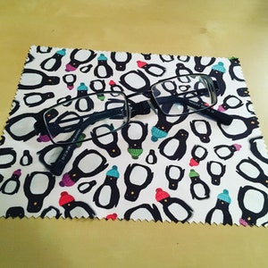 May include: A white microfiber cleaning cloth with a black border and a repeating pattern of penguins wearing colorful hats. A pair of black eyeglasses is lying on top of the cloth.