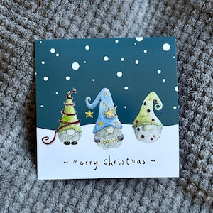 May include: A square Christmas card with three gnome illustrations. Each gnome wears a pointed hat, one green, one blue, and one green. The card has a dark blue background with white snowflakes and the words "merry christmas".
