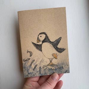 May include: A brown card featuring a black and white illustration of a puffin with a yellow beak. The puffin is standing on a rock with small fish swimming around it.