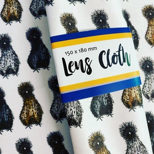 May include: White fabric with a repeating pattern of black and brown chickens. The fabric has a blue and yellow label that says "Lens Cloth 150 x 180 mm".