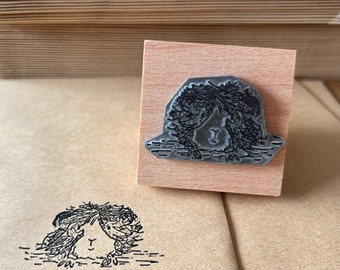 May include: A wooden block stamp with a black and white illustration of a guinea pig with its head peeking out from under a pile of hay. The stamp is on a wooden base.