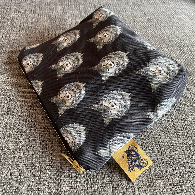 Puede incluir: Bolsa de lona negra con cremallera con un patr&oacute;n repetido de caras de lobos grises. La bolsa tiene una cremallera dorada y una peque&ntilde;a etiqueta de cuero marr&oacute;n con un dise&ntilde;o de bicicleta.