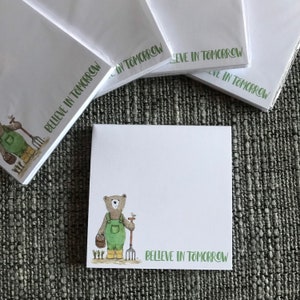 Peut inclure: Une pile de blocs-notes blancs avec une bordure verte et le texte "Believe in Tomorrow" imprimé sur chaque bloc-notes. Le bloc-notes du dessus présente une illustration de dessin animé d'un ours brun portant une salopette et tenant un outil de jardinage.